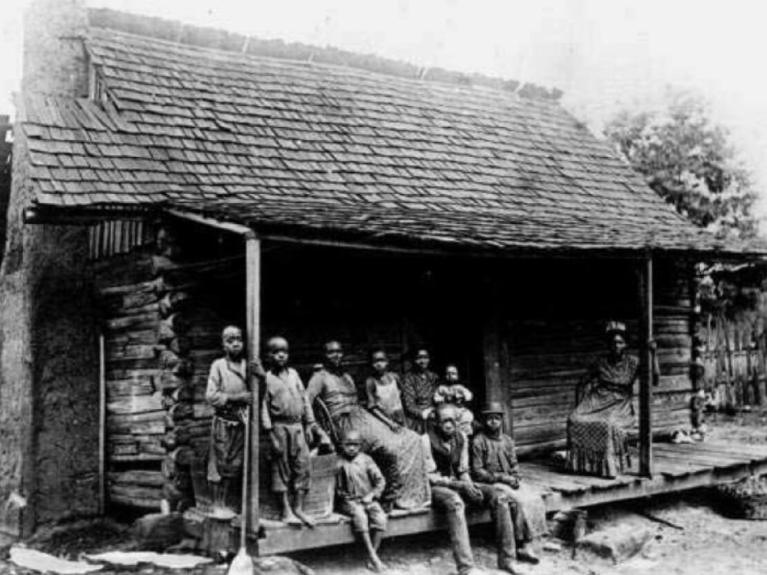 A black and white photo showing a group of people standing and sitting on the wooden porch of a house.