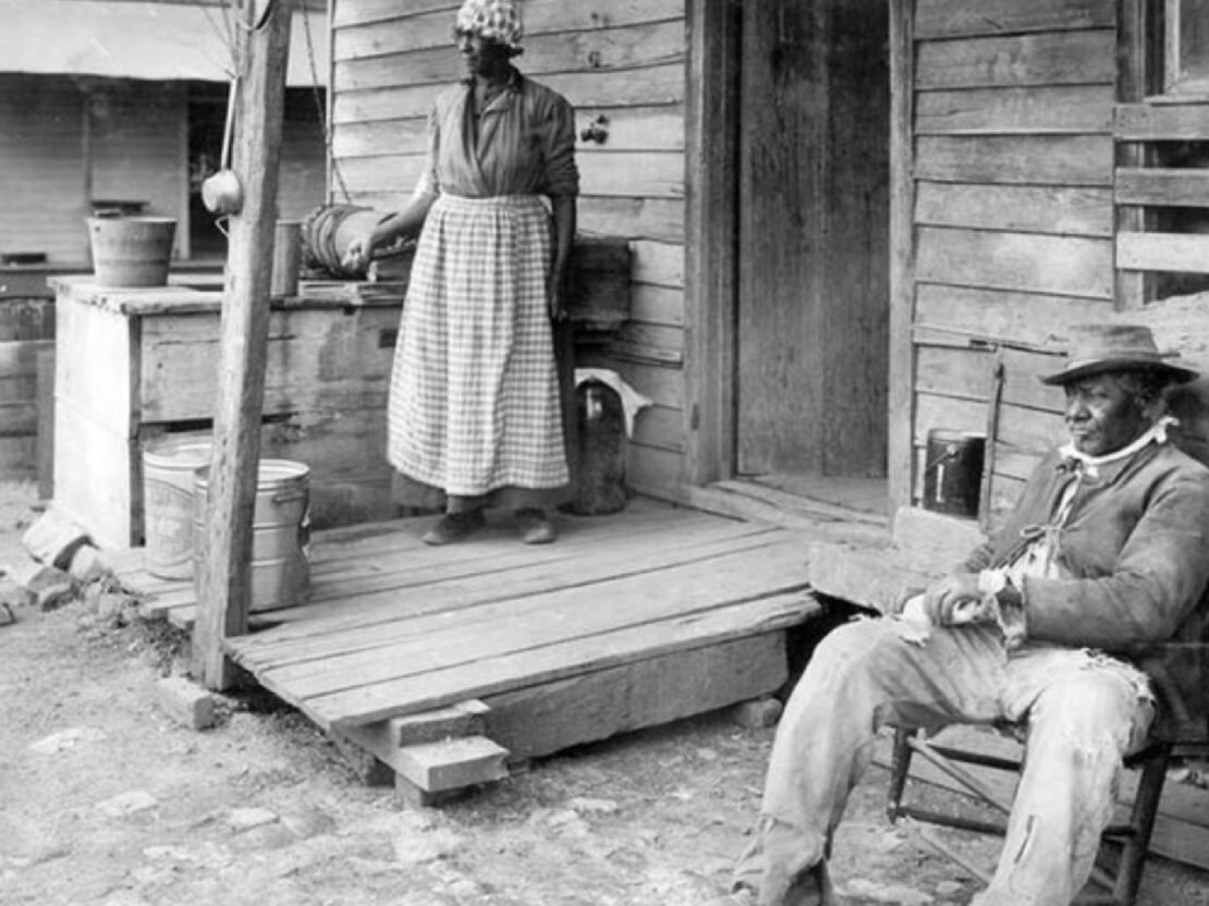 A black and white image of a man seated on a chair outside a wooden cabin. A woman stands on a porch to the left of him.