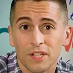 Man in a plaid shirt speaking in front of a wall with colorful handwritten text.