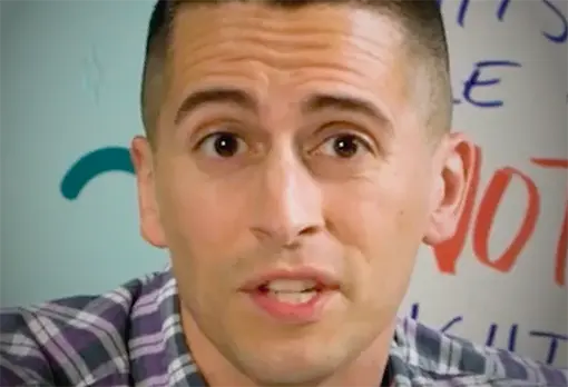 Man in a plaid shirt speaking in front of a wall with colorful handwritten text.