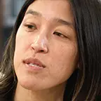 Woman with straight black hair looking to the side, speaking in a library setting.