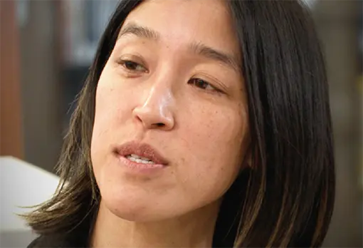 Woman with straight black hair looking to the side, speaking in a library setting.