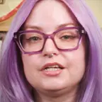 Woman with long lavender hair and purple glasses speaking in front of library bookshelves.