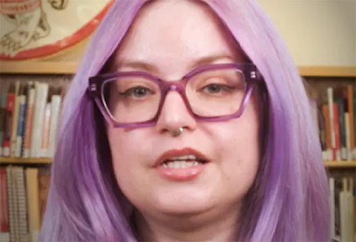 Woman with long lavender hair and purple glasses speaking in front of library bookshelves.