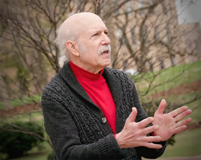 Man with a bald head and gray mustache gesturing with both hands while speaking outdoors in a red turtleneck and black cardigan.