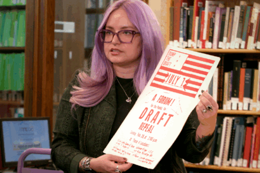 Person with long lavender hair holding a red-and-white protest flyer reading “Dump the Draft – July 1 – A Forum for the Public to DRAFT Repeal.”