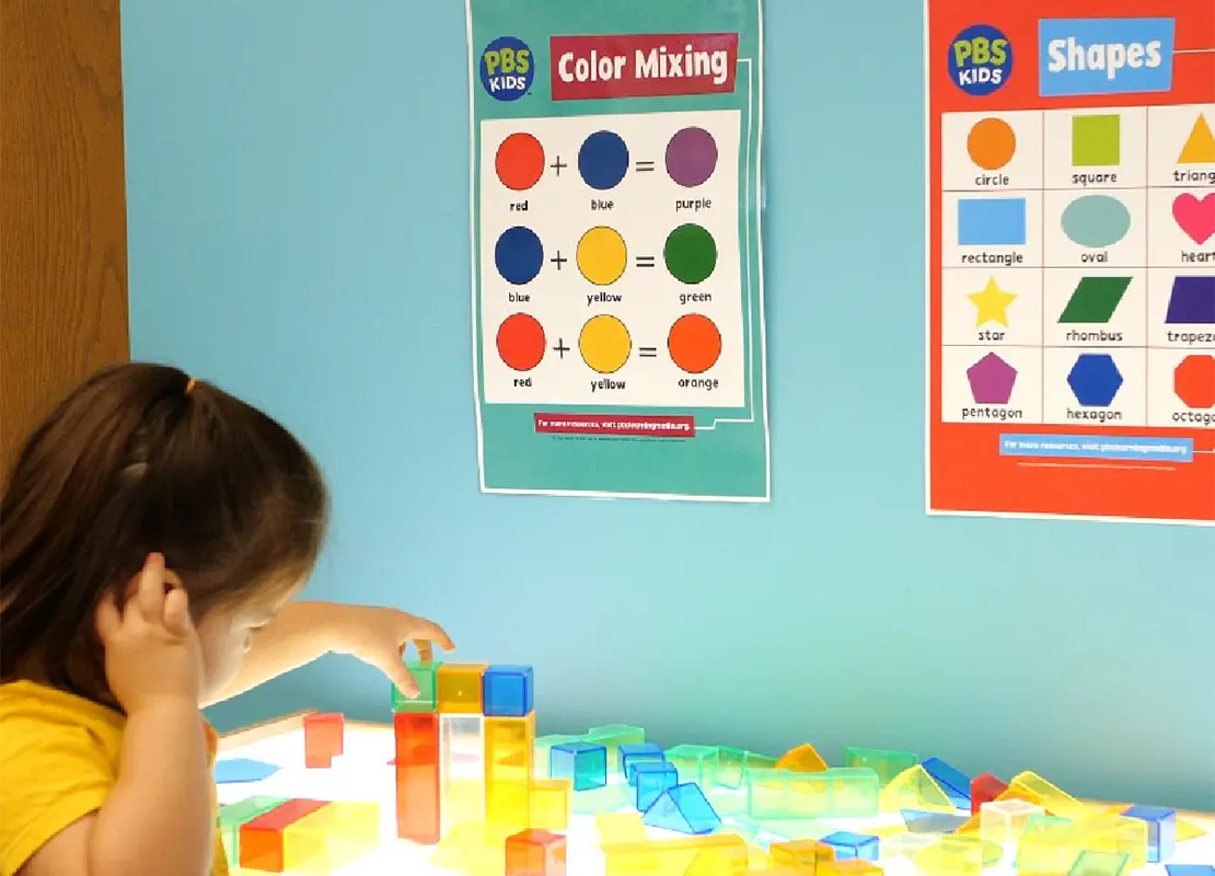 Child playing with colored blocks with PBS KIDS posters in the background that teach about colors and shapes