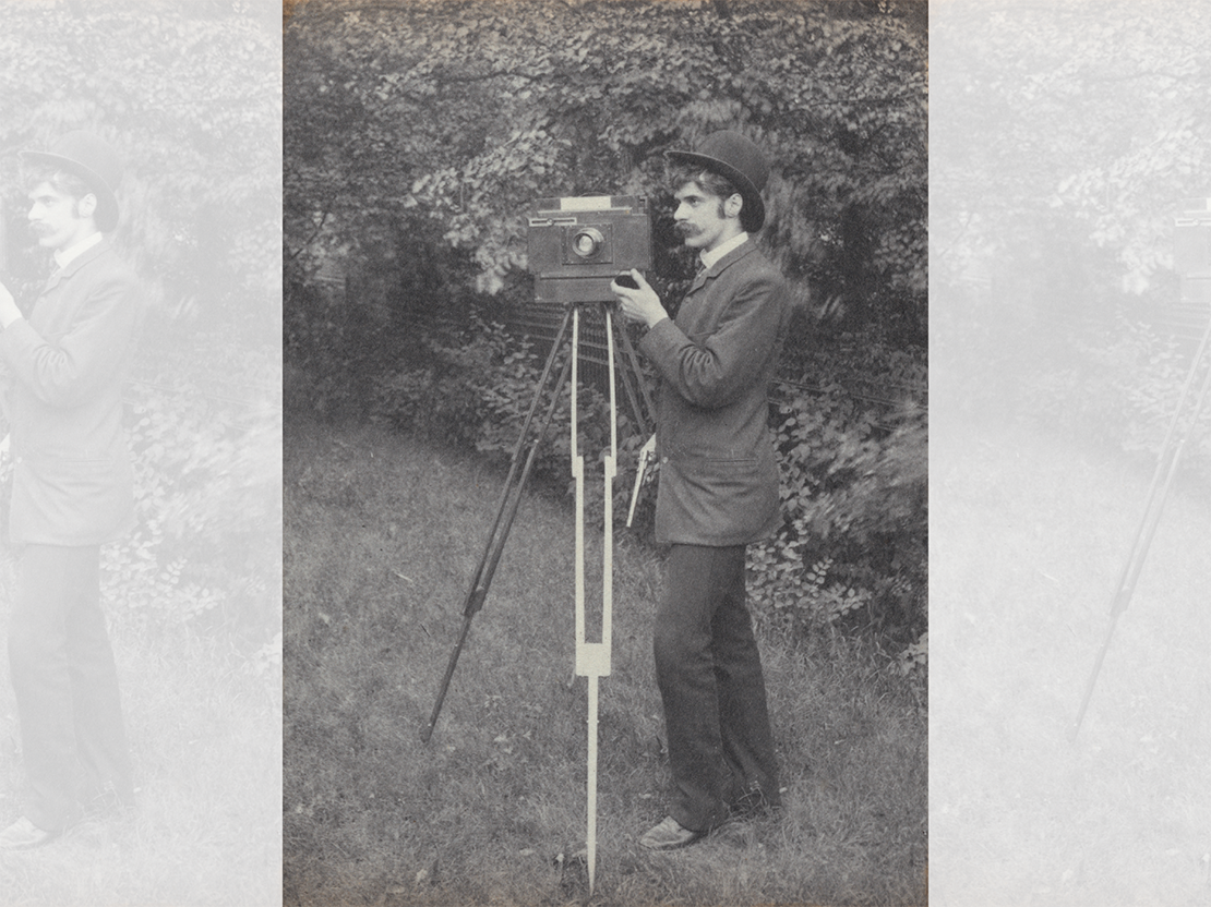 A black and white photograph of a young man standing outside in a suit and hat taking a picture with an old-fashioned camera.