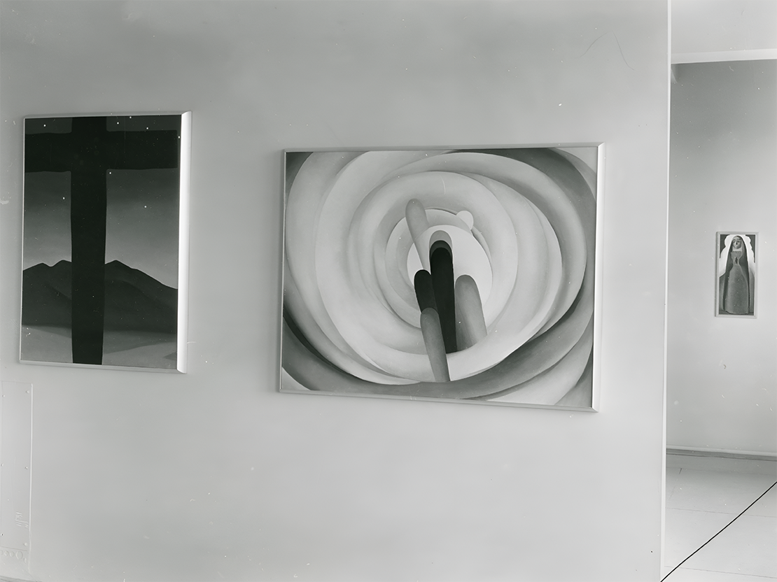 A black and white photo showing three of Georgia's paintings, two of which are on a wall in the foreground. The main painting shows a close up of a flower.