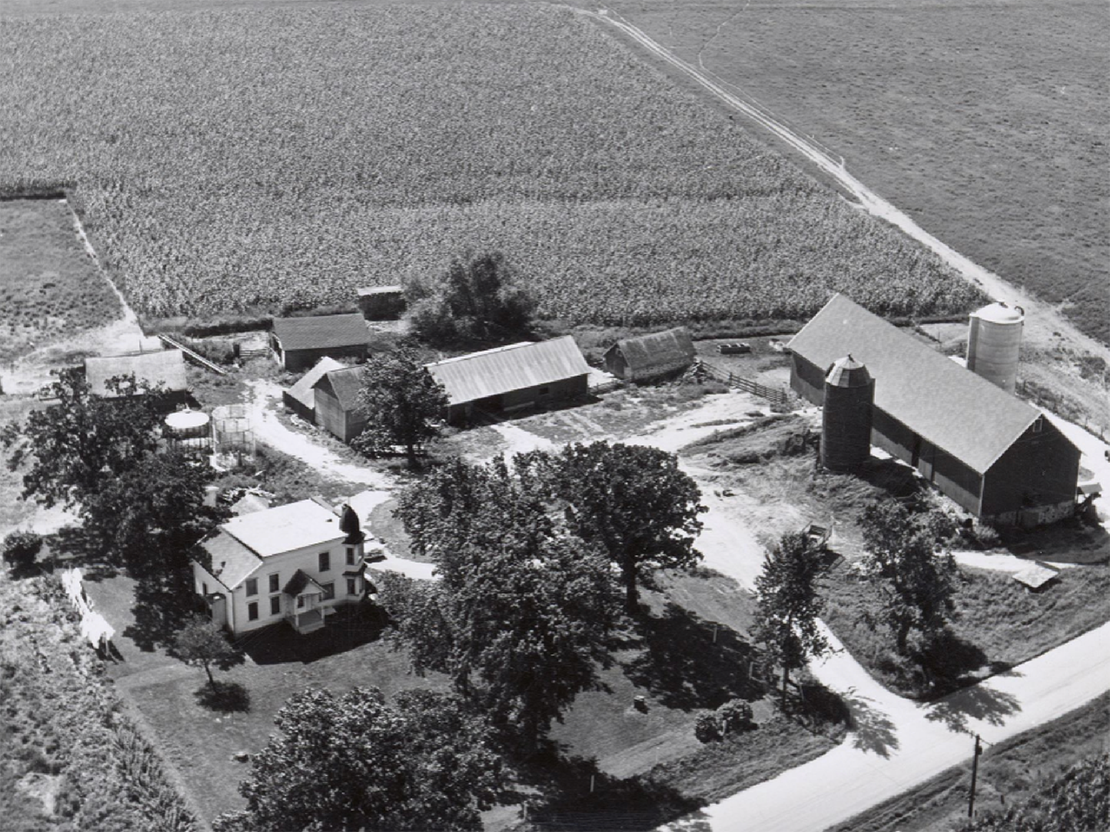 A black and white aerial photo of a farm, showing fields, trees, a house, and several barns and smaller buildings.