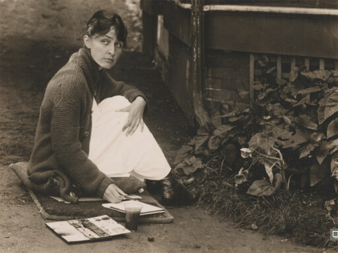A black and white photograph of Georgia O'Keeffe sitting on a mat on the ground with her painting supplies.