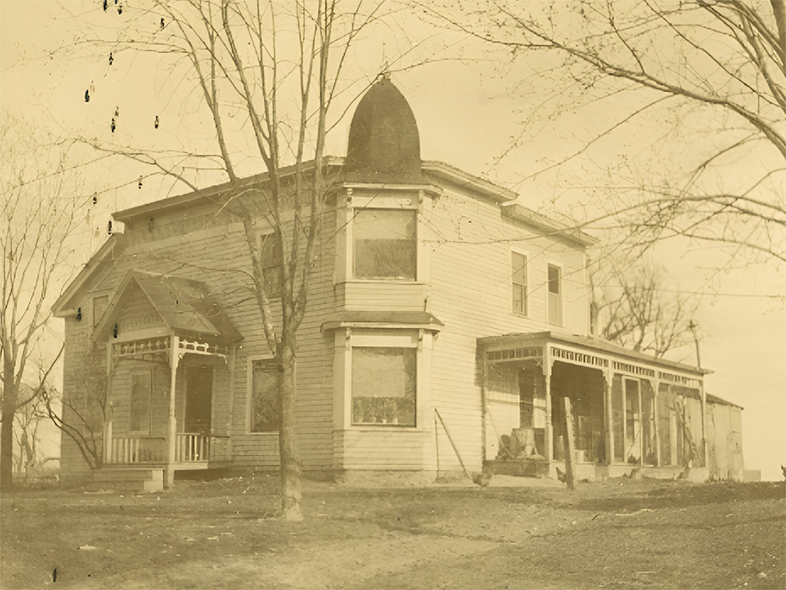 A black and white photograph of a house with wood siding, a small porch entrance, and a large porch on the other side.