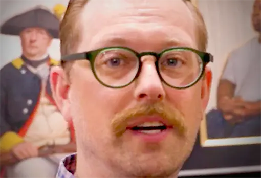 Man with reddish-blond hair, mustache, and round glasses speaking in front of a historical portrait.