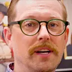 Man with reddish-blond hair, mustache, and round glasses speaking in front of a historical portrait.