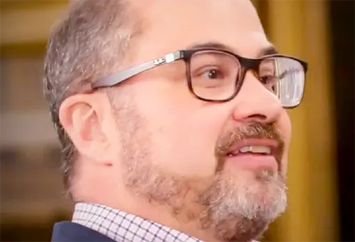 Man with a trimmed beard and rectangular glasses smiling while speaking, golden frame in background.