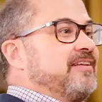 Man with a trimmed beard and rectangular glasses smiling while speaking, golden frame in background.