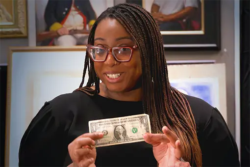 Woman with glasses and braided hair smiling while holding a U.S. one-dollar bill in front of framed portraits.