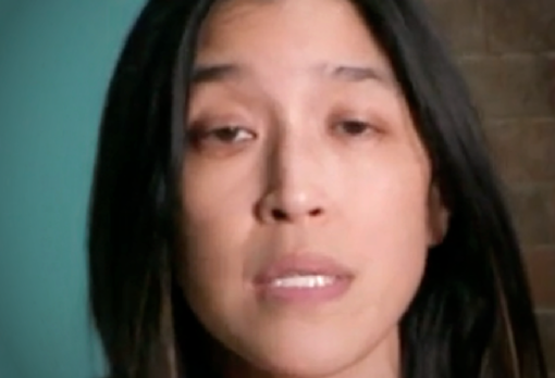 Woman with long straight hair and neutral expression speaking in front of a teal wall and brick background.
