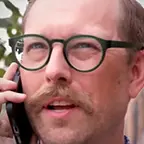 Man with a mustache and green round glasses talking on a phone outside, with trees in the background.