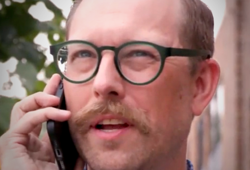Man with a mustache and green round glasses talking on a phone outside, with trees in the background.