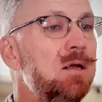 Man with a styled mustache, beard, and glasses speaking indoors with light from tall windows behind him.