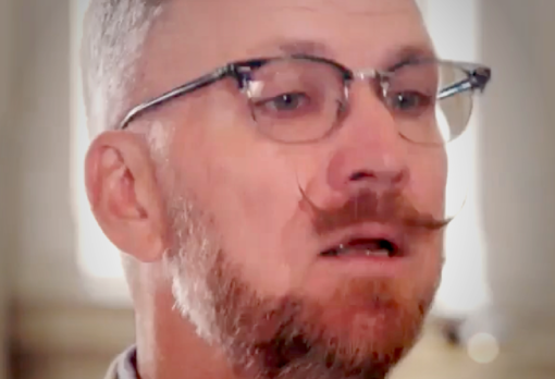 Man with a styled mustache, beard, and glasses speaking indoors with light from tall windows behind him.