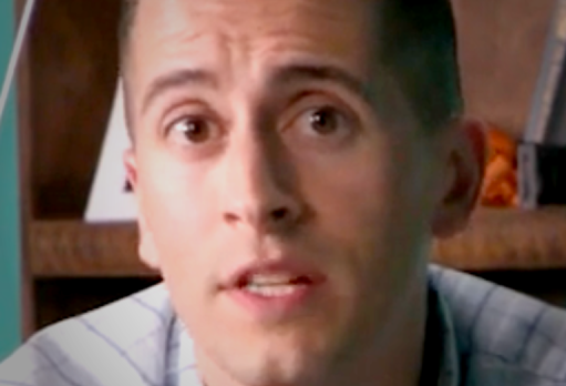 Young man in a checkered shirt speaking in front of bookshelves in a studio setting.