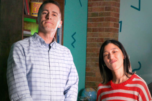 Two hosts standing in front of a teal wall with abstract shapes, one in a plaid shirt and one in a red-striped sweater.