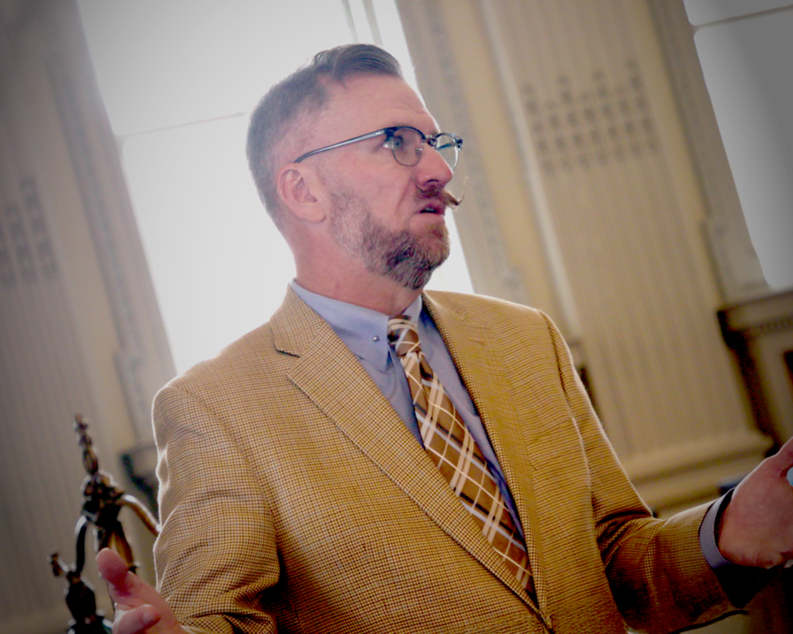 Man with glasses and a neatly trimmed beard wearing a tan blazer speaking in a historic interior with tall windows.