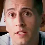 Young man in a checkered shirt speaking in front of bookshelves in a studio setting.