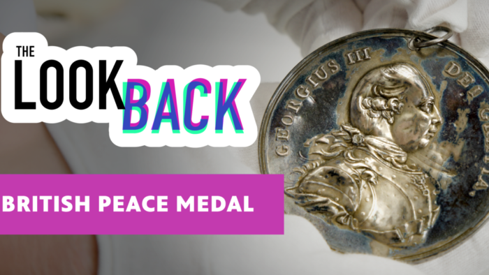 A close-up of a silver British peace medal depicting King George III in profile, held with white gloves. Text overlay reads “The Look Back” and “British Peace Medal.”