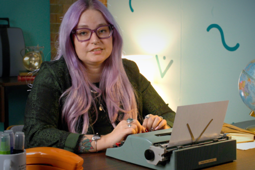 Person with long lavender hair and purple glasses at a desk with a vintage typewriter, looking at the camera.