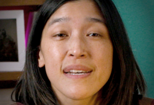 Close-up of person with straight dark hair speaking, with teal wall and bookshelf in background.