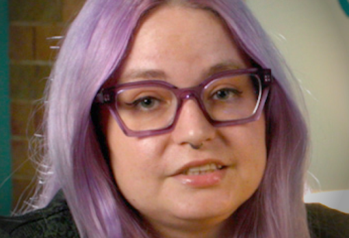 Person with short hair wearing purple glasses and a patterned sweater, looking slightly left.