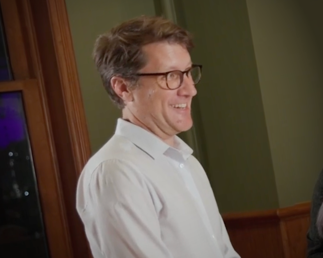 Man in white shirt and glasses smiling in profile near a window, in a warmly lit interior.