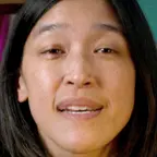 Close-up of person with straight dark hair speaking, with teal wall and bookshelf in background.