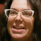 Person with long dark hair and white cat-eye glasses speaking, with a red curtain background.