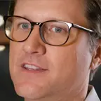 Close-up of man in dark-rimmed glasses and white shirt, looking slightly right in a softly lit room.