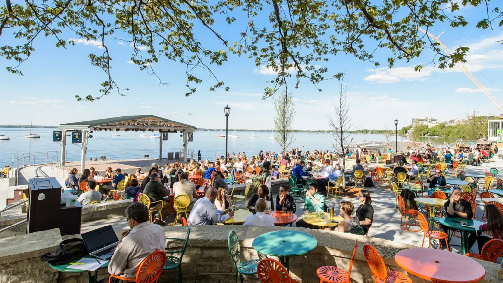 Memorial Terrace at University of Wisconsin–Madison on a sunny day with visitors seated at the Union's trademark colorful metal tables and chairs overlooking Lake Mendota