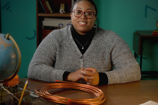 Host seated at a desk with a coil of copper tubing and a globe, smiling at camera in front of a teal wall