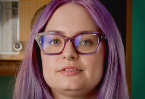 Person with purple hair and glasses speaking to camera in a studio setting