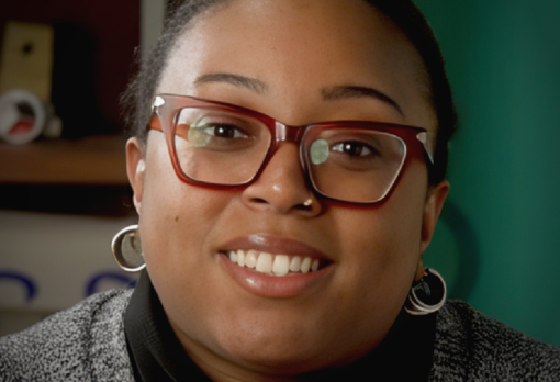 Host smiling warmly at camera, wearing hoop earrings and glasses with a bookshelf behind her