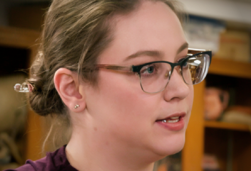 Person with glasses and hair pulled back speaking in profile, museum display cases visible behind