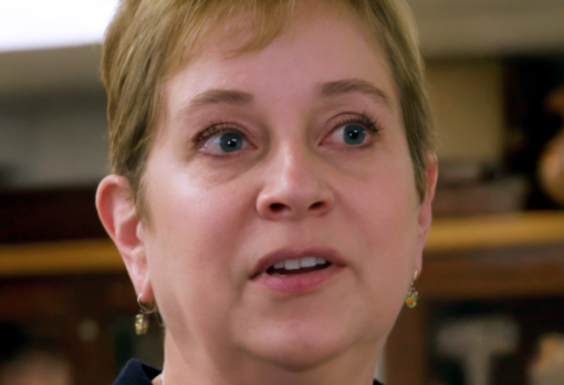 Person with short hair and gold earrings speaking to camera with display cases in background