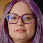 Person with purple hair and glasses speaking to camera in a studio setting