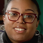 Host smiling warmly at camera, wearing hoop earrings and glasses with a bookshelf behind her