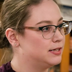 Person with glasses and hair pulled back speaking in profile, museum display cases visible behind