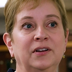 Person with short hair and gold earrings speaking to camera with display cases in background