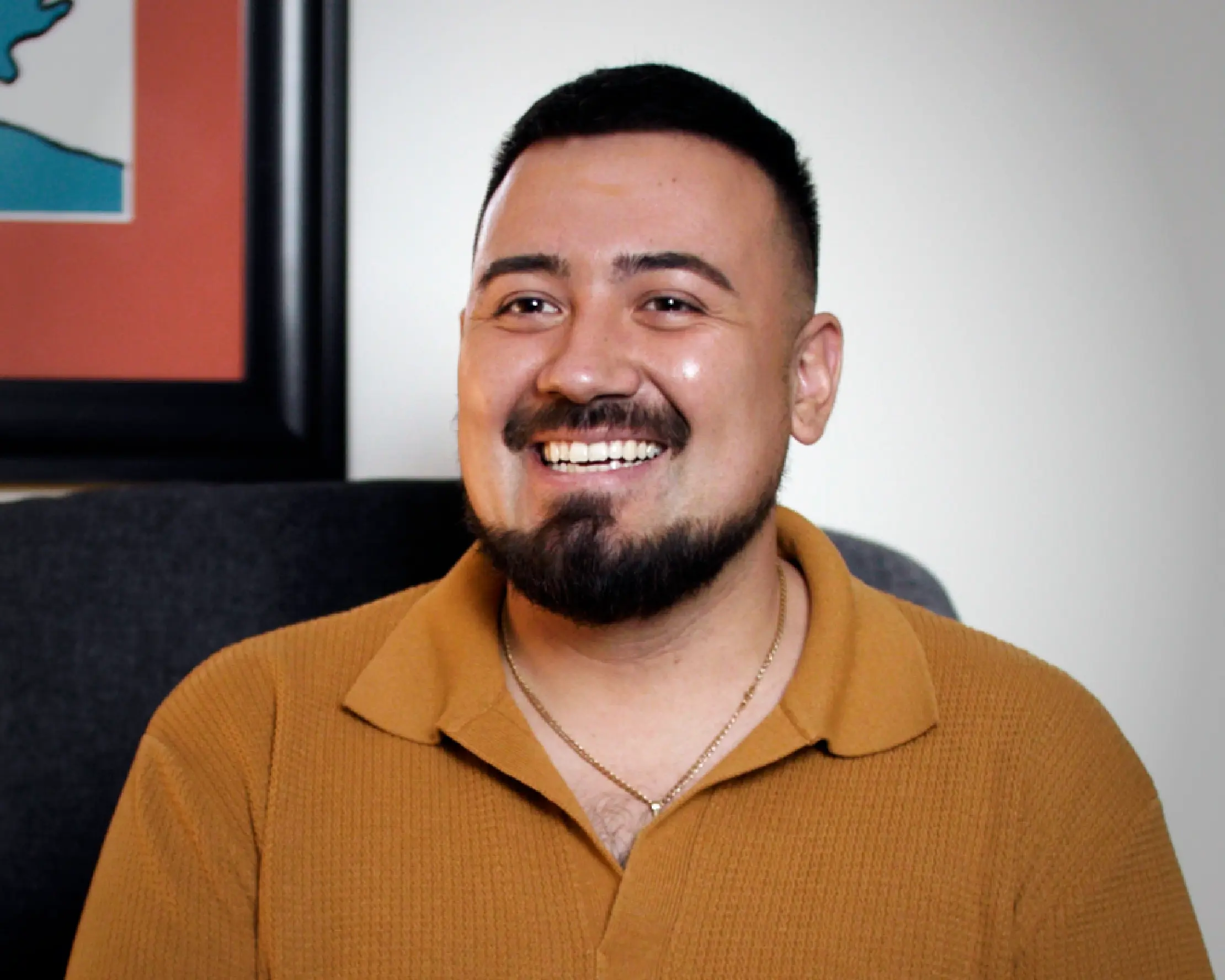 A young man with a beard smiles warmly, wearing a mustard yellow polo shirt and a gold chain necklace. A framed artwork with orange and teal tones is visible in the background.