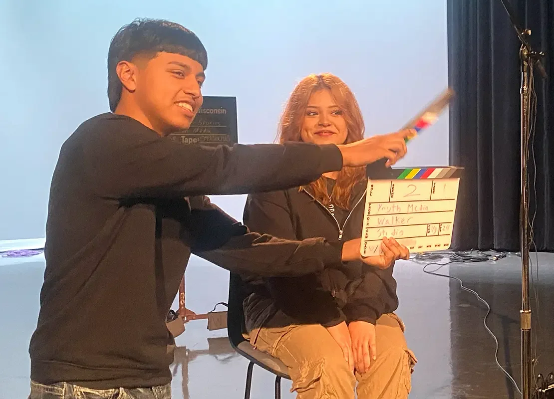 A person operating a clapperboard in front of a person who is being interviewed. Both people are smiling.